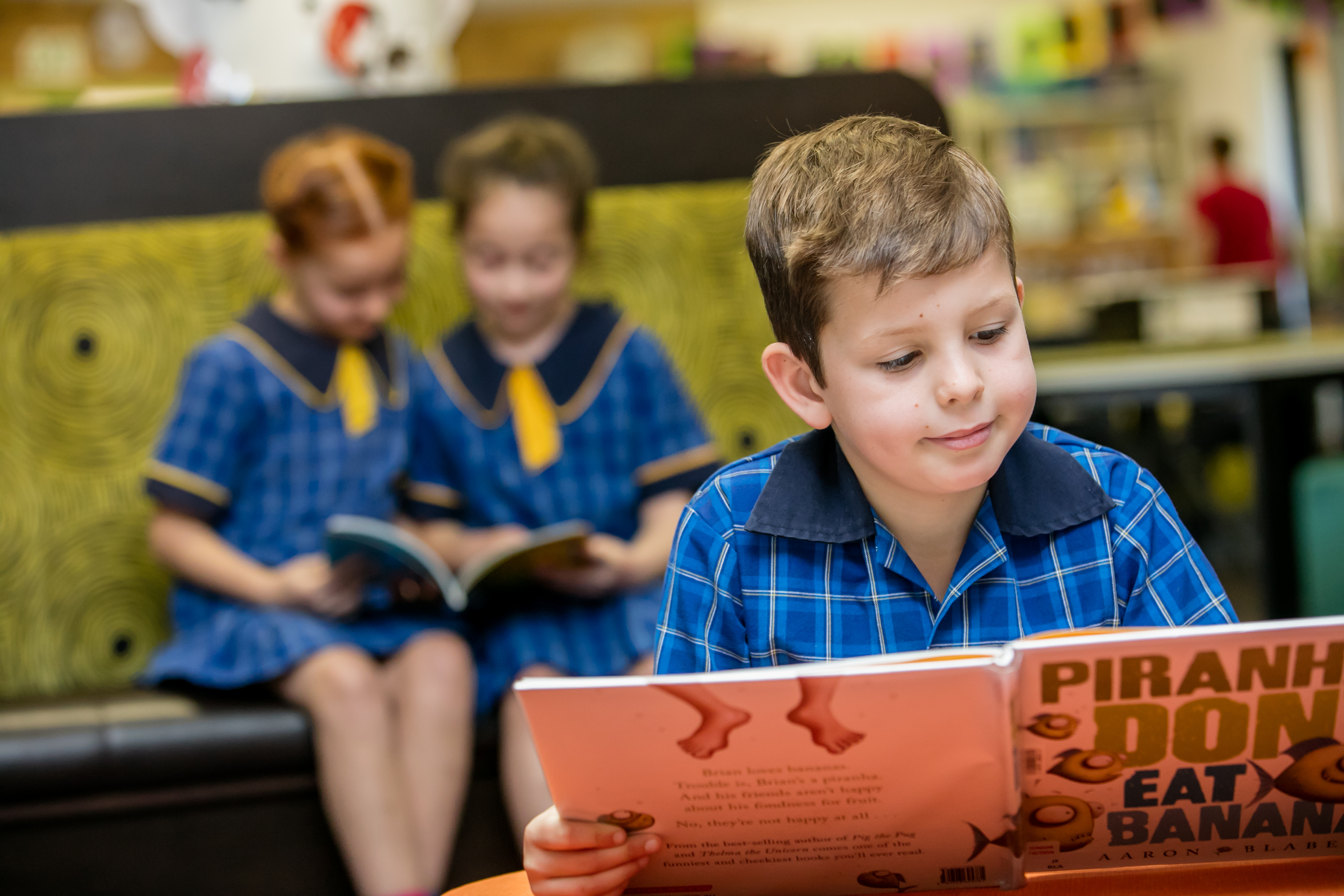 Boy reading book in the library. In the background there are two girls sitting on the couch and reading. 
